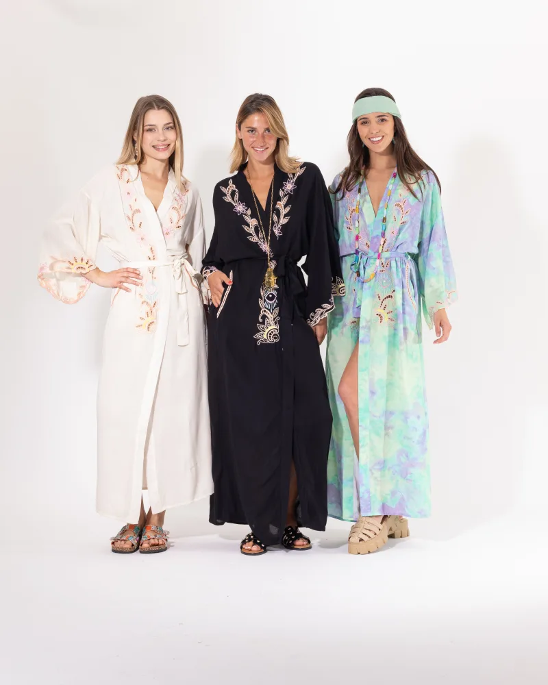 Trois femmes souriantes portant des kimonos longs bohèmes à broderies florales et motifs ajourés, déclinés en trois coloris : écru, noir et tie-dye vert/violet, ceinturés à la taille.